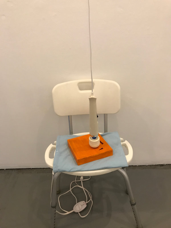 Image Description: A white shower chair with silver metallic legs placed in front of a wall. Seated on it is a light blue heating pad, with a small wood board painted orange with two deep red geometric shapes. On top of it is a white headed handheld massager hanging from the ceiling, slightly caressing the surface of the wood board.