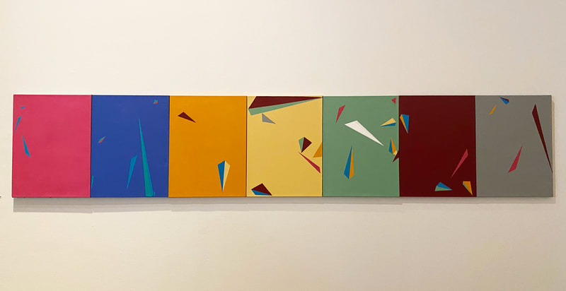 seven canvases are hung immediately next to each other, forming one large string of paintings. they all have a variety of triangles painted on them, and from left to right, are: pink, blue, orange, yellow, sage green, maroon, and grey.

