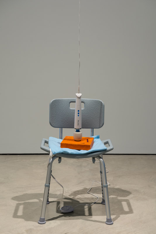 Image Description: A grey shower chair with silver metallic legs placed in front of a wall. Seated on it is a light blue heating pad, with a small wood board painted orange with two deep red geometric shapes. On top of it is a white headed handheld massager hanging from the ceiling, slightly caressing the surface of the wood board.