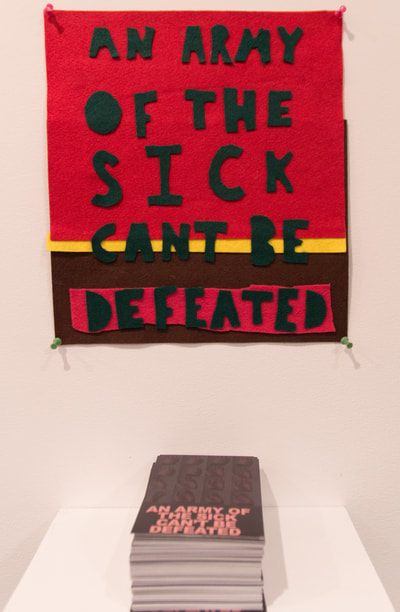 felt banner with text in green that says an army of the sick can’t be defeated, on a red background that is on the top 3/4ths of the image, with a yellow stripe then brown, and a pink strip on top of the brown. below is a stack of postcards with pink text that reads an army of the sick can't be defeated.