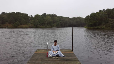 Ezra is wearing a light blue hospital gown, staring directly to the camera. He is centered in the photograph sitting cross legged on a wooden dock, with trees and a lake as the backdrop on a grey skied day, giving an ominous and contemplative ambiance.