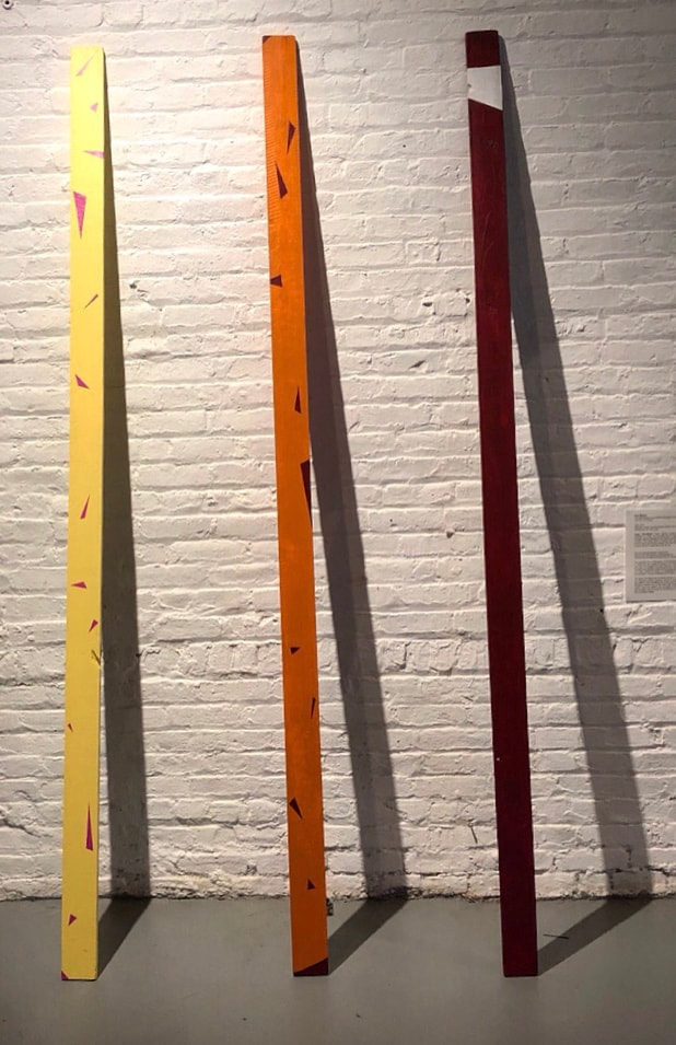 hree painted wooden planks leaning and centered on a wall. The left most plank is light yellow with magenta triangles painted throughout the surface seemingly floating, falling, or crashing. The center plank is orange with deep red triangles on the surface seemingly floating, falling, and crashing. The right most plank is deep red with a white band near the top. Shadows casted from each plank make three large triangular structures